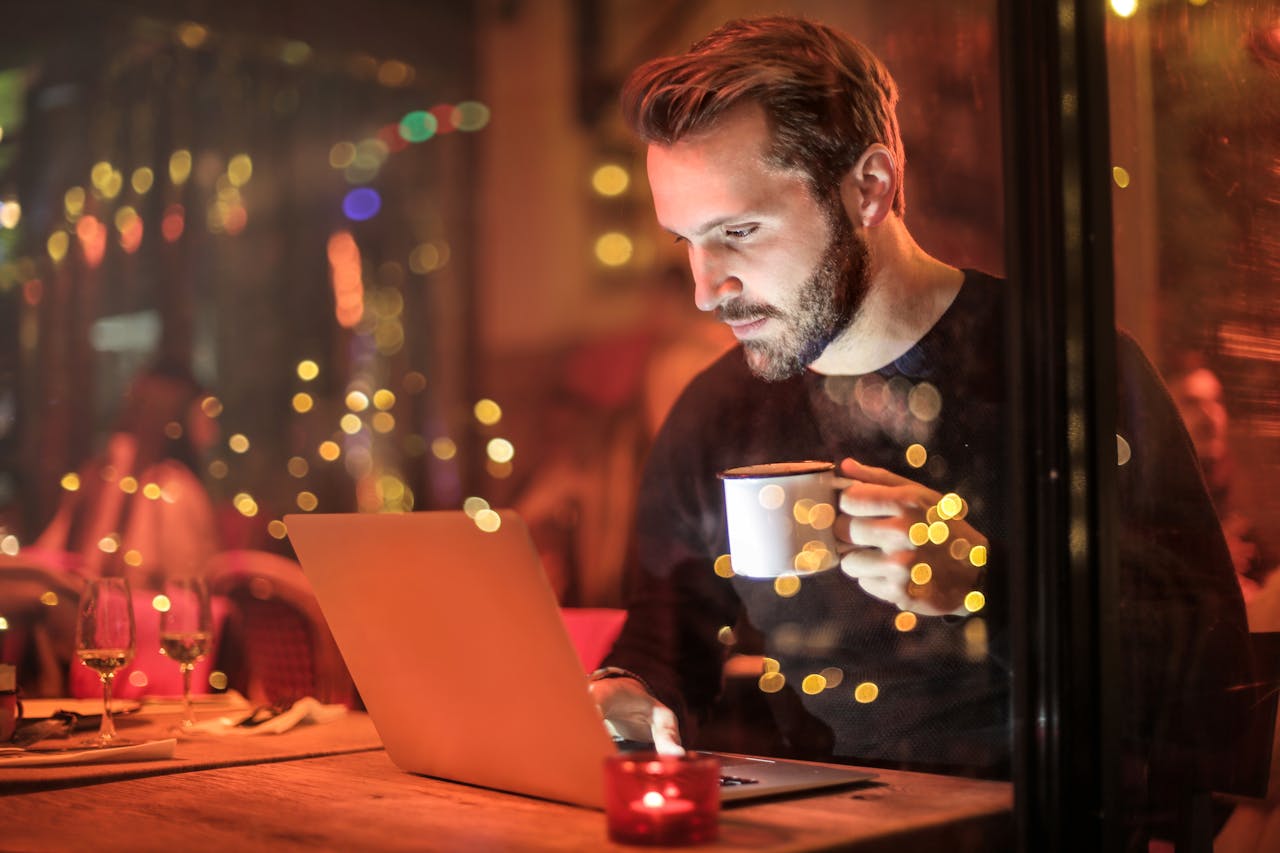 Home A young man with a beard is working on a laptop at a cozy cafe with warm lighting, holding a coffee cup.